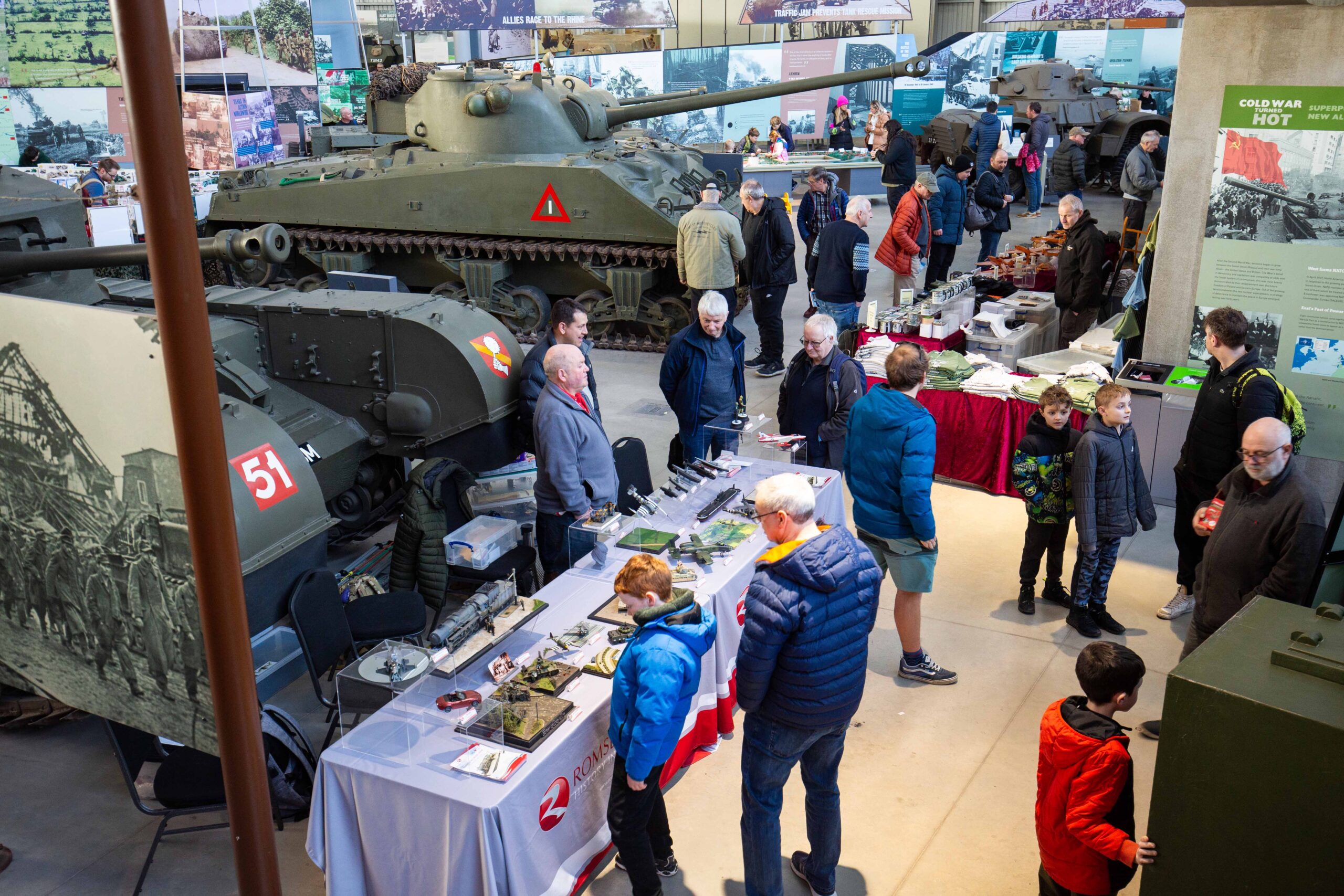 Model Show February 2025_004 Visitors in exhibition looking at scale models on tables in front of tanks.