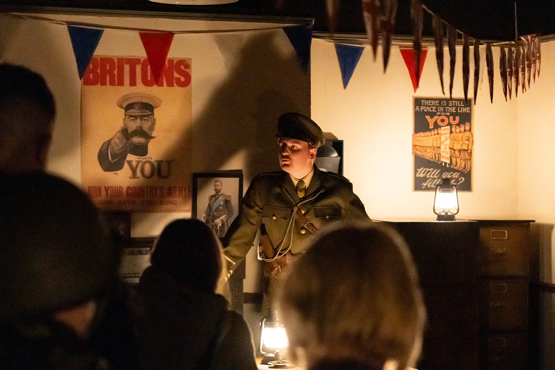 Night at The Tank Museum - 1 Man in solider's uniform stood in front of bunting and WWI posters, lit by lamplight.