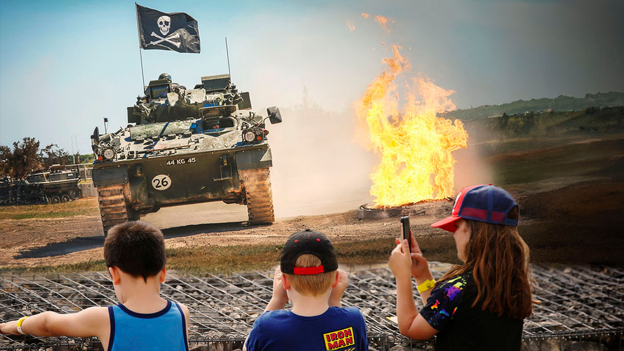 3 children watching an armoured vehicle drive around the arena with an explosion